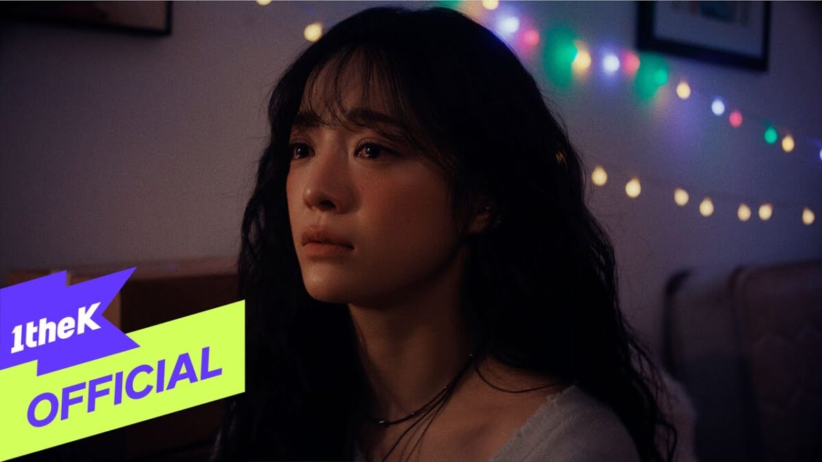 Kim Sejeong com expressão emotiva, iluminada por luzes coloridas em um ambiente aconchegante.